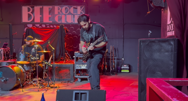 Second Illusion guitarist playing electric guitar during a live performance at BFE Rock Club, with drummer performing in the background under warm lighting.