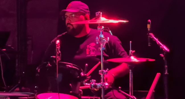 Second Illusion drummer performing live on stage under red lighting, focused mid-performance behind the drum kit at BFE Rock Club.
